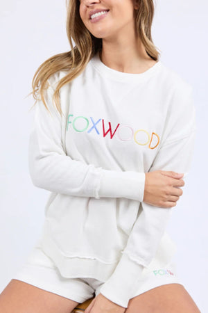 A woman sits on a stool, smiling with her arms loosely crossed, wearing the Foxwood Simplified Confetti Crew—a white 100% cotton sweatshirt and shorts set with "FOXWOOD" in multicoloured embroidery across the front.