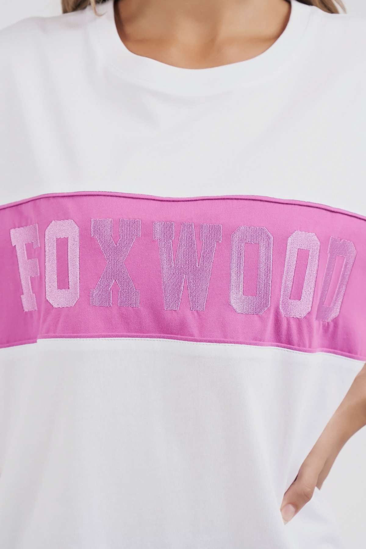 A person models the Foxwood Connection Tee, a white cotton shirt with a pink stripe and "FOXWOOD" embroidered in pink letters, offering casual leisurewear vibes as their hand rests on their hip.