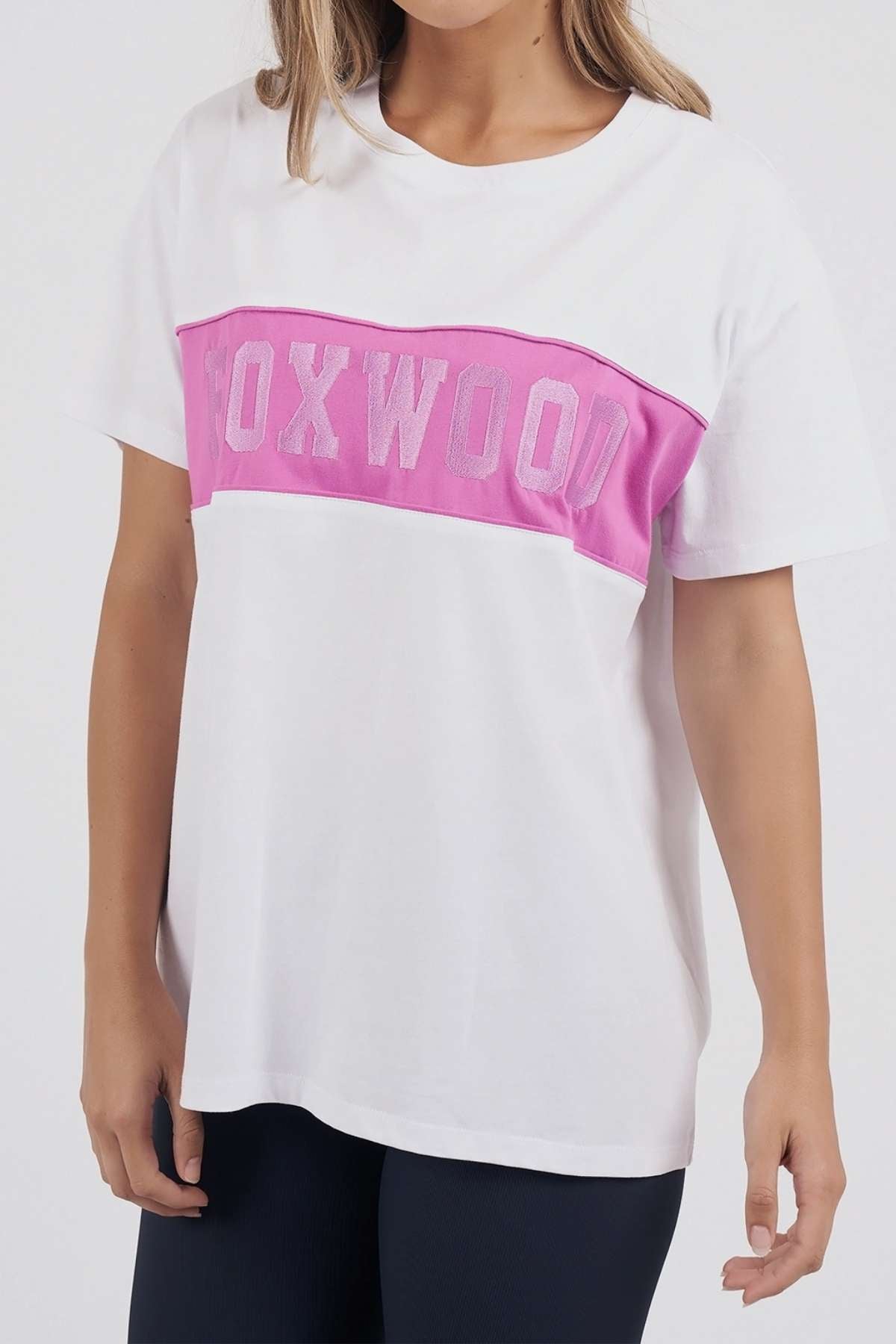Wearing the Foxwood Connection Tee, a leisurewear t-shirt with a pink stripe and "FOXWOOD" across the chest, made from 100% cotton. It pairs well with dark pants against a plain white background.