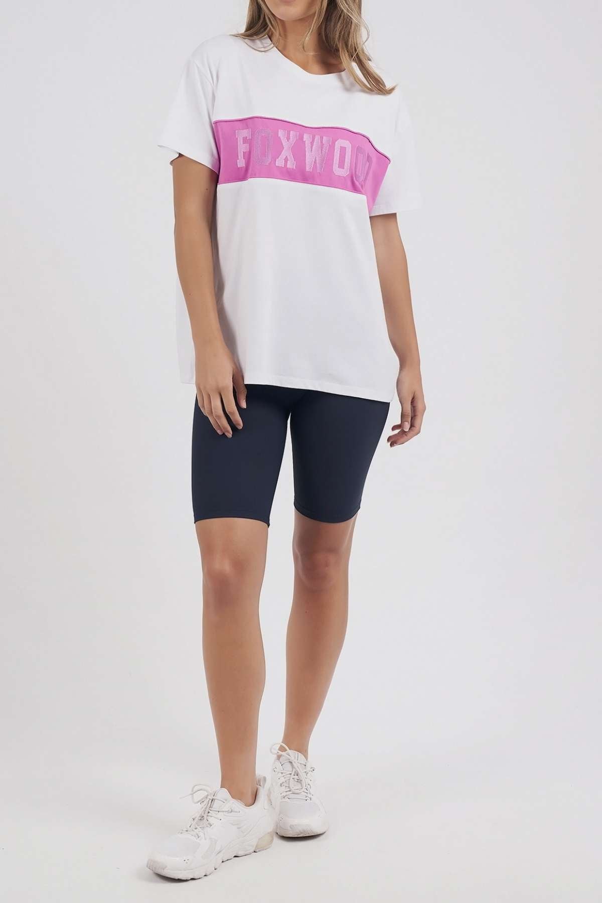 A person showcases the Foxwood Connection Tee, a 100% cotton white T-shirt with a pink "FOXWOOD" banner, paired with black cycling shorts and white sneakers against a simple backdrop, exemplifying stylish leisurewear.