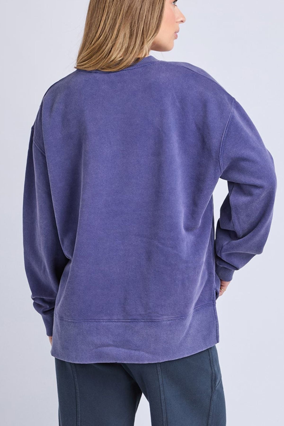 A person with light brown hair wears the oversized Foxwood Core Crew—a loose, long-sleeve purple cotton sweatshirt—and dark pants, shown from the back against a plain light background.
