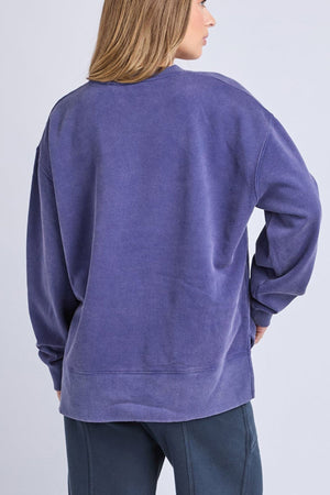 A person with light brown hair wears the oversized Foxwood Core Crew—a loose, long-sleeve purple cotton sweatshirt—and dark pants, shown from the back against a plain light background.