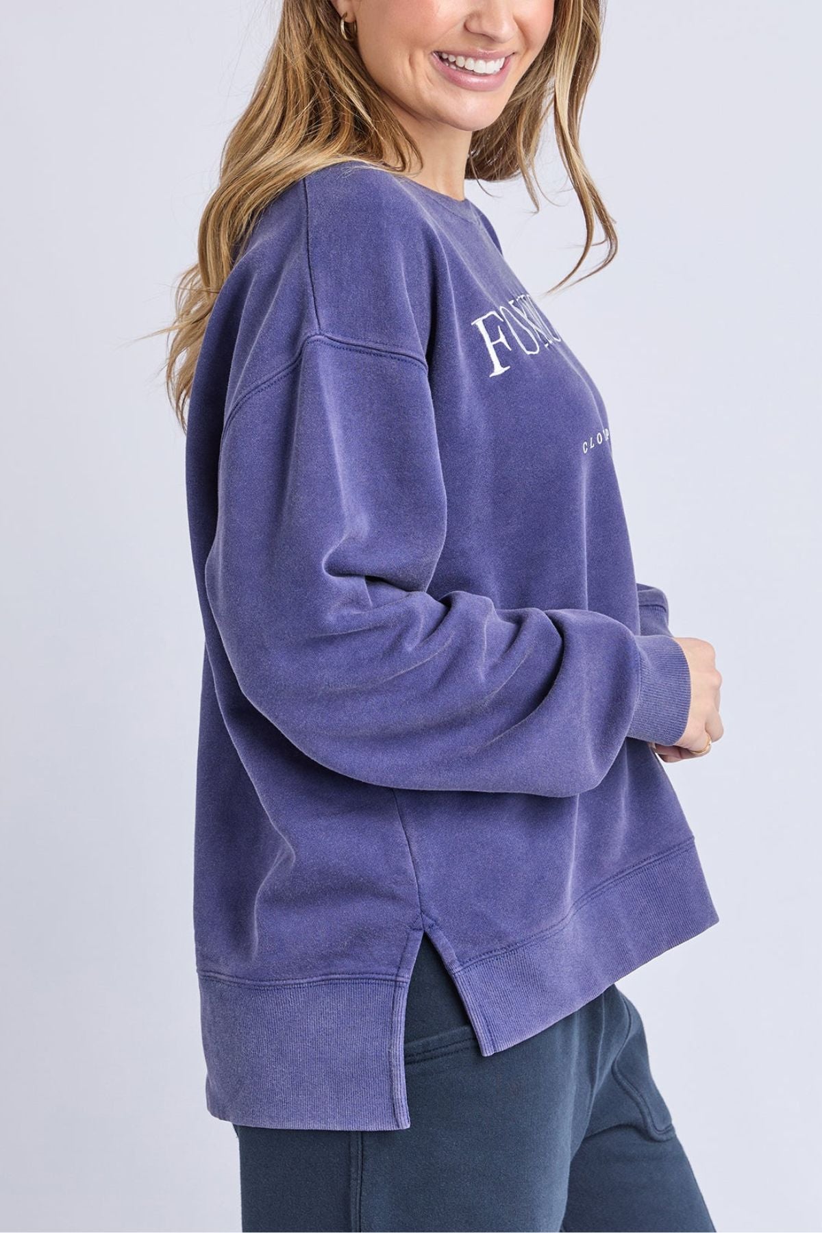 A woman with long hair smiles wearing the Foxwood Core Crew, an oversized purple cotton sweatshirt with side slits, relaxed fit, dark pants, and white text plus Foxwood artwork on the chest.