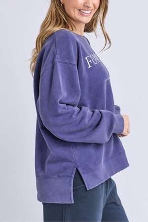 A woman with long hair smiles wearing the Foxwood Core Crew, an oversized purple cotton sweatshirt with side slits, relaxed fit, dark pants, and white text plus Foxwood artwork on the chest.