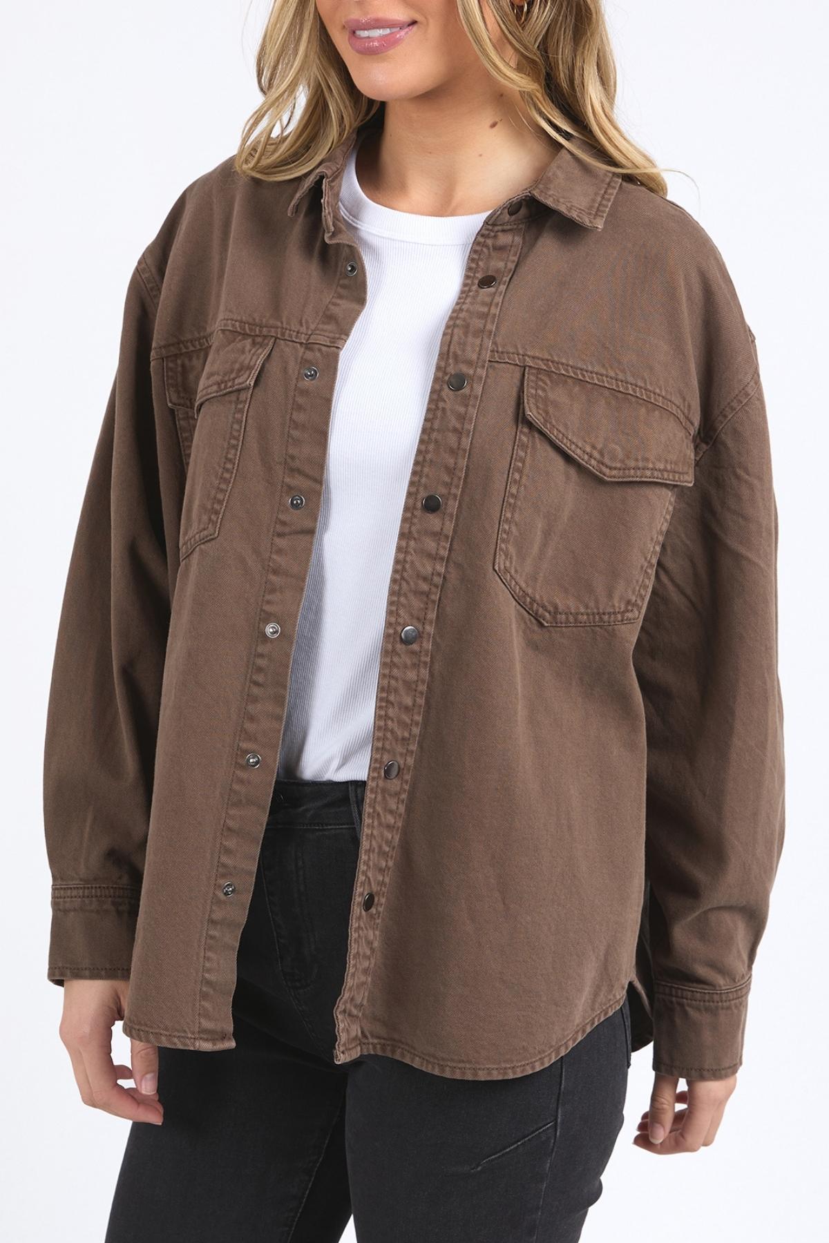 A woman wears the Foxwood Daisy Overshirt, a relaxed-fit brown cotton shirt with a button-through placket, layered over a white t-shirt and black pants, standing against a plain white background.