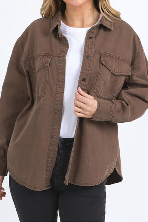 A person wears the Foxwood Daisy Overshirt—a relaxed fit brown cotton shirt with a button-through placket—over a white top and black pants, standing against a plain white background. Only the upper body is visible.