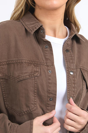 A woman wears a white ribbed cotton shirt and the Foxwood Daisy Overshirt, an unbuttoned brown denim jacket in a relaxed fit, holding its front with both hands. Only her lower face and torso are visible.