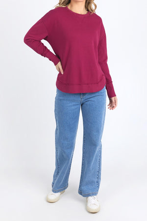 A person wearing a long-sleeved maroon Foxwood Delilah Crew, light blue jeans, and white sneakers stands with one hand on their hip against a plain white background.