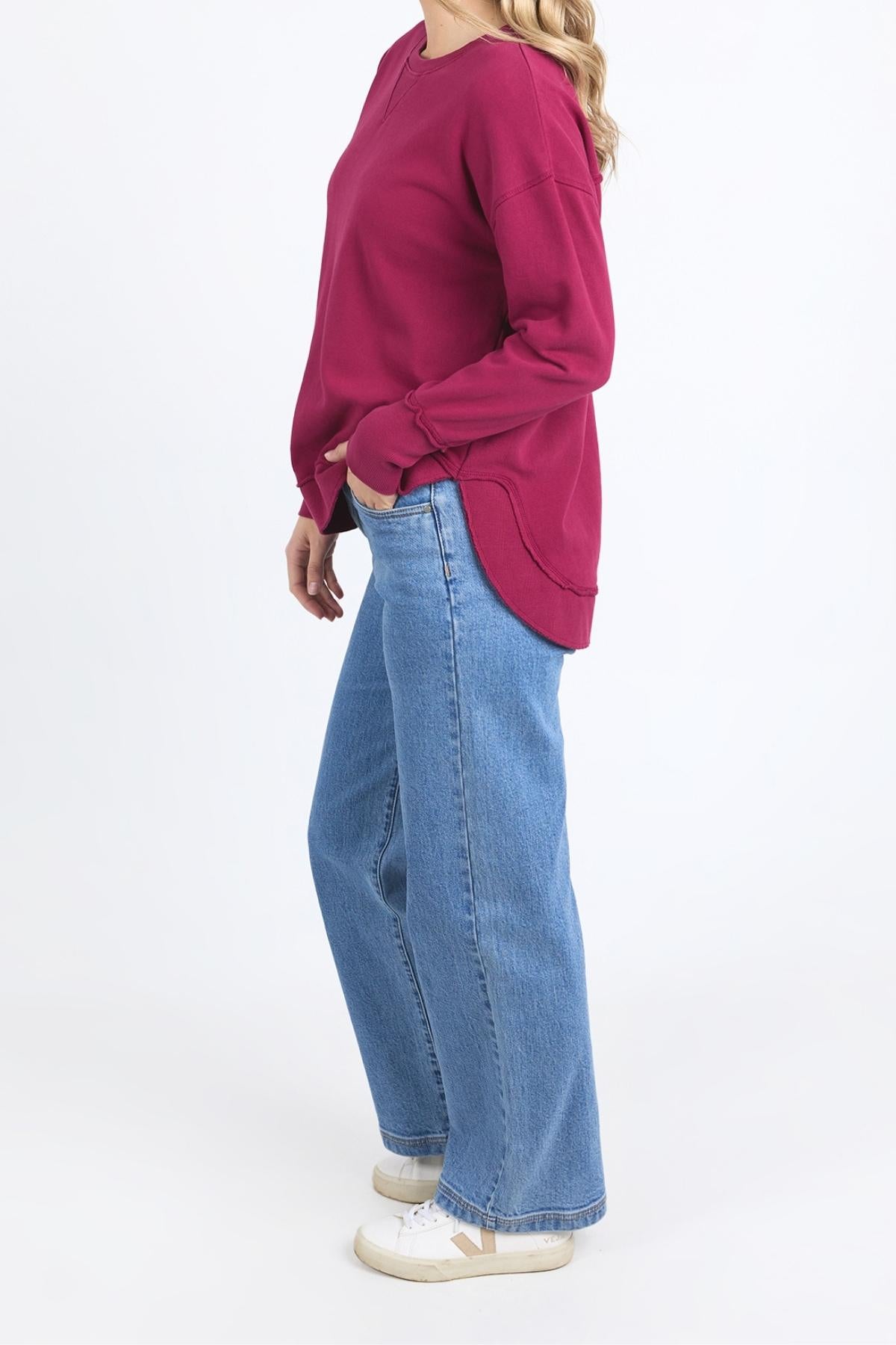 A person stands sideways wearing a maroon Foxwood Delilah Crew, loose blue jeans, and white sneakers against a plain white background.