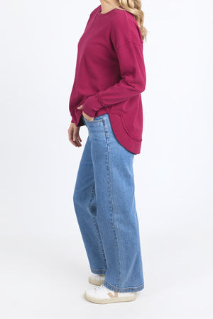 A person stands sideways wearing a maroon Foxwood Delilah Crew, loose blue jeans, and white sneakers against a plain white background.