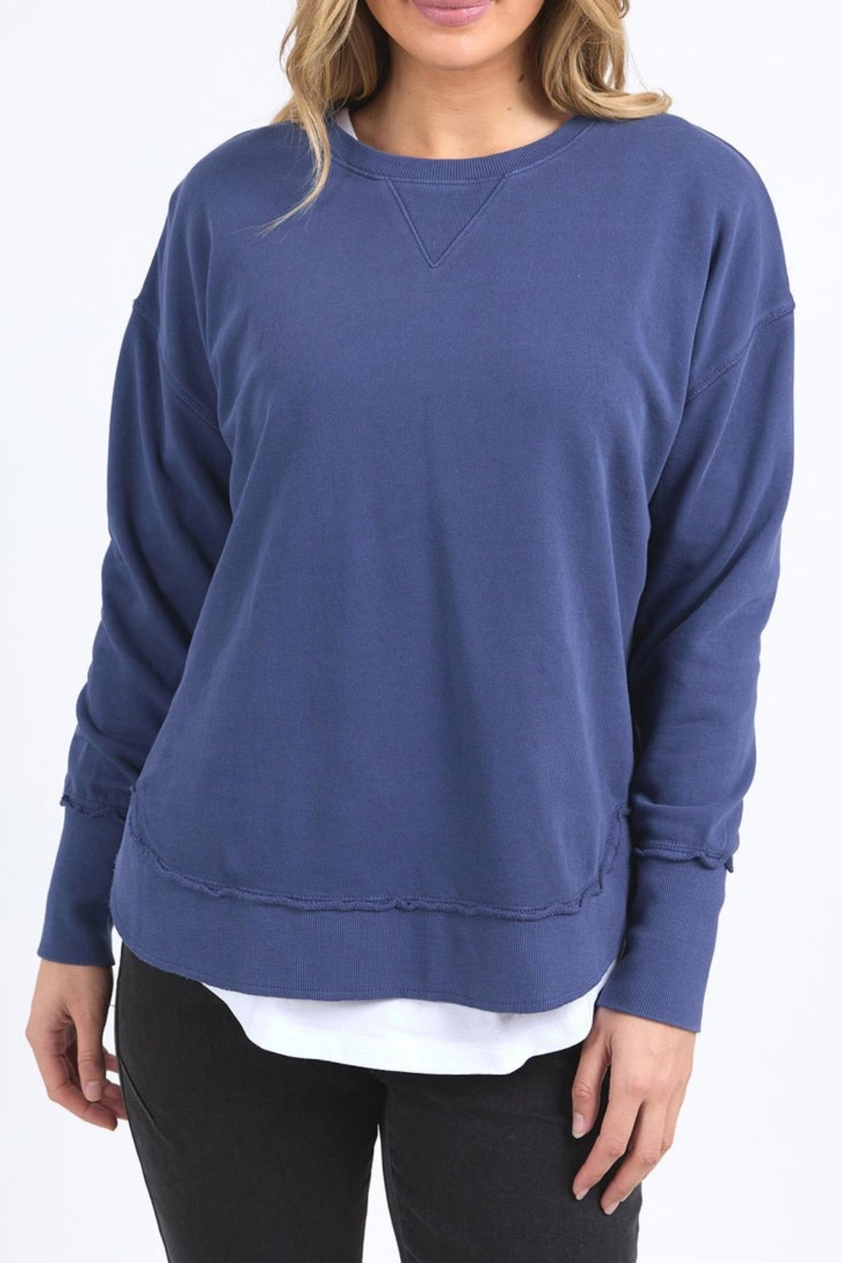 A person wearing the Foxwood Delilah Crew in blue over a white shirt with black pants stands against a plain white background. The face is partially visible.