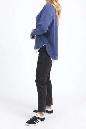 A woman stands sideways wearing the blue Foxwood Delilah Crew, black jeans, and black Adidas sneakers with white soles. Her long blonde hair is down against a plain white background.
