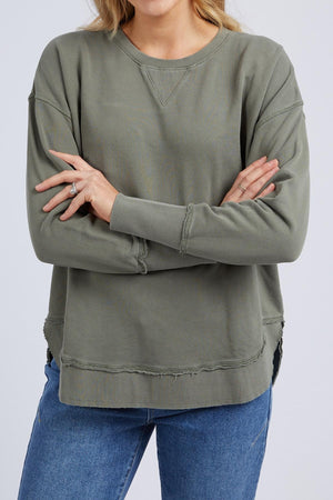 A woman with long blonde hair wears the Foxwood Delilah Crew, a loose olive green cotton jumper with raw edges, paired with blue jeans. She stands with arms crossed in front of her torso; her face is partially out of frame.