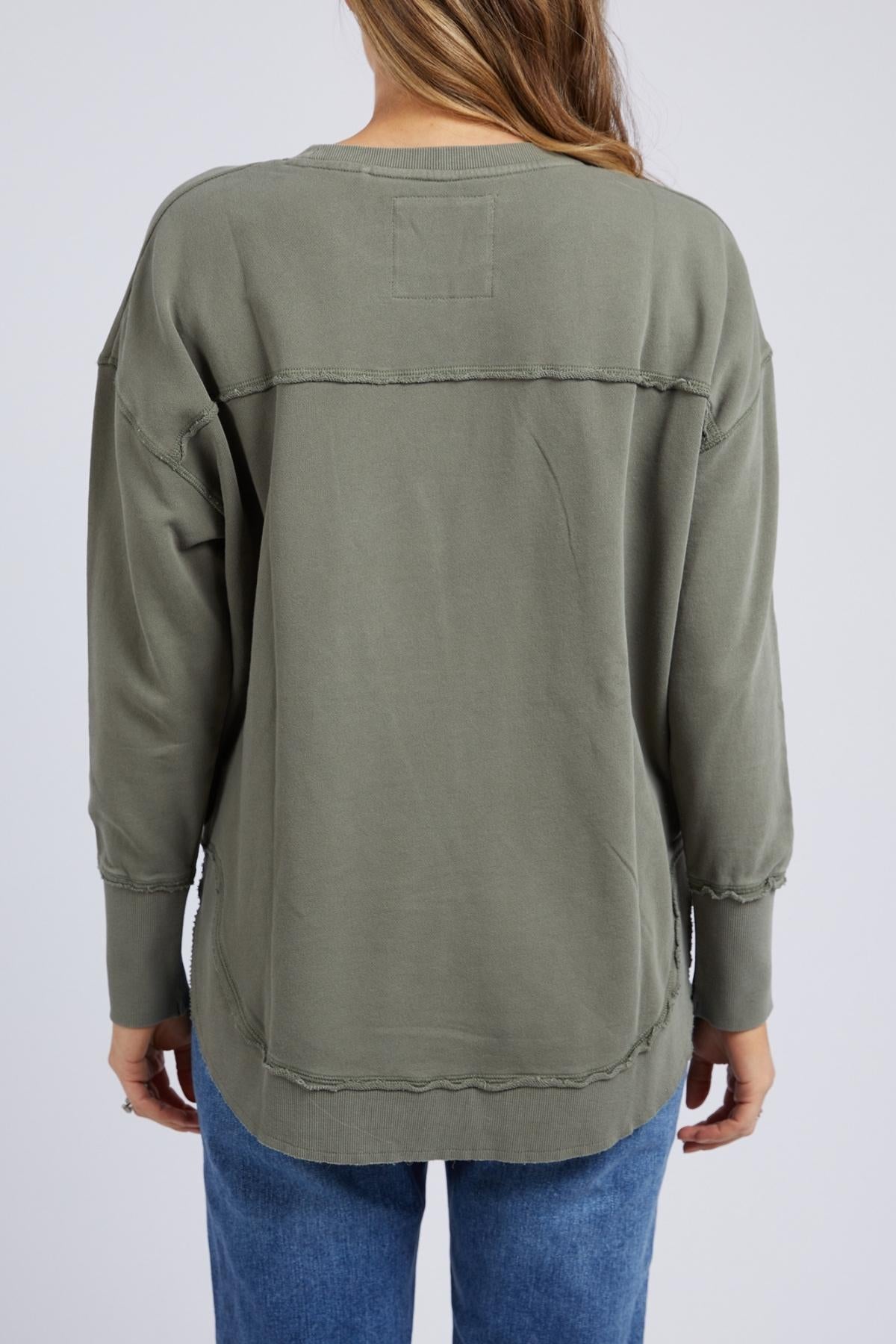 A person with long hair, shown from the back, wears the Foxwood Delilah Crew—an olive green, loose-fitting cotton jumper with visible seams—paired with blue jeans against a plain light background.