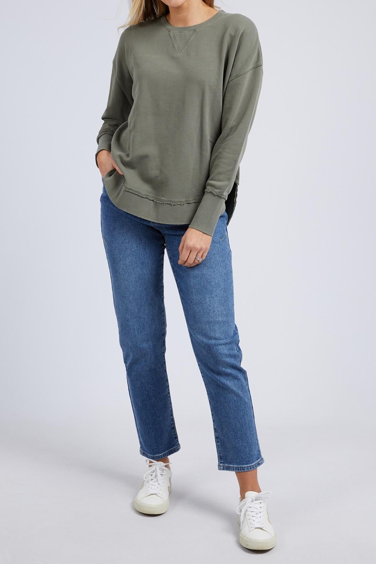 A person stands against a plain background wearing the Foxwood Delilah Crew jumper in olive green, blue jeans, and white sneakers, with one hand in their pocket and the other touching their hair. Their head is not visible in the image.