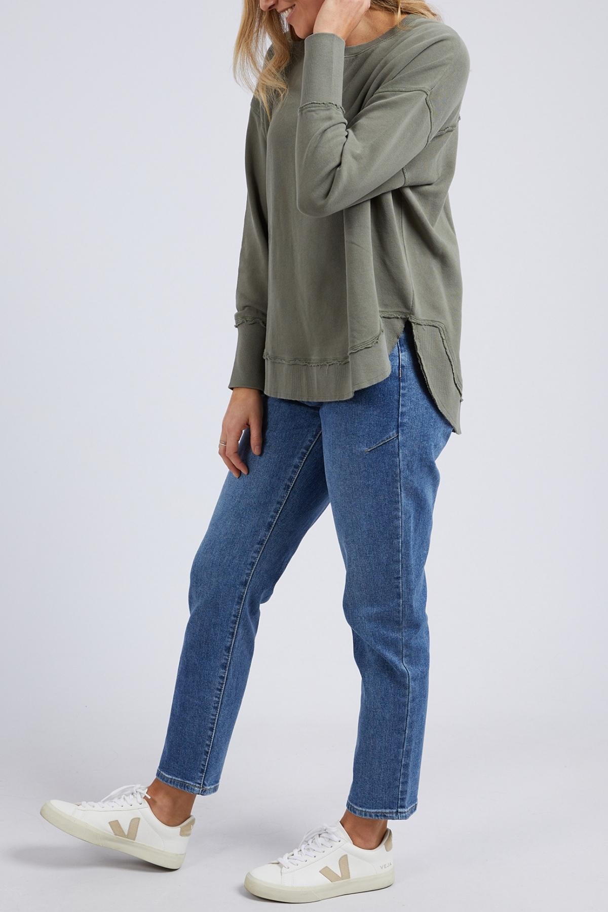A person with light brown hair, head partially out of frame, stands against a plain background wearing the Foxwood Delilah Crew in olive green, blue jeans, and white sneakers with beige details.