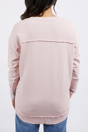 A person with long dark hair wears a light pink Foxwood Delilah Crew and blue jeans, standing with their back to the camera against a plain white background.