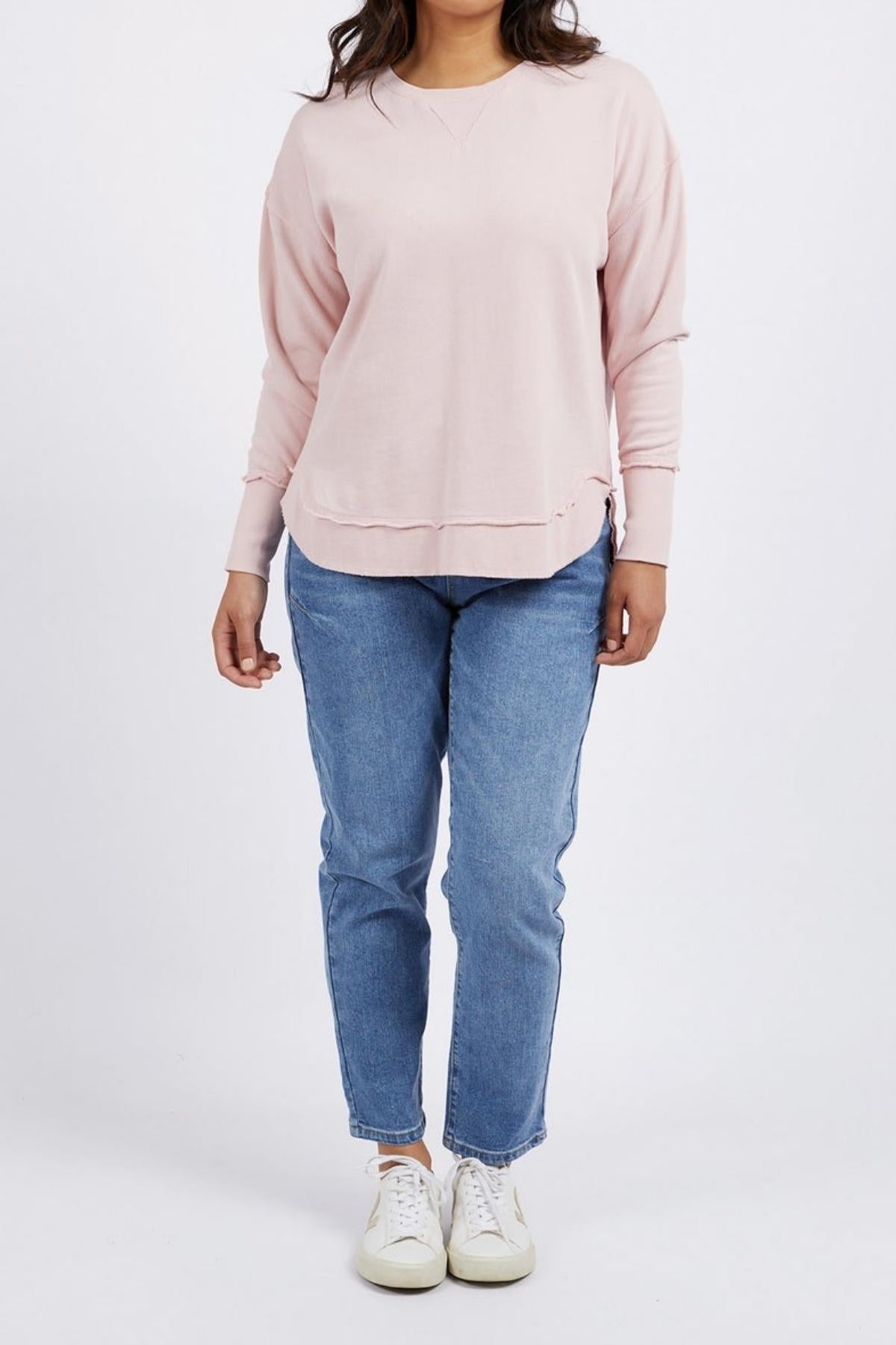 A person stands facing forward, wearing a light pink Foxwood Delilah Crew, blue jeans, and white sneakers. The background is plain white, with the person’s head partially out of frame.