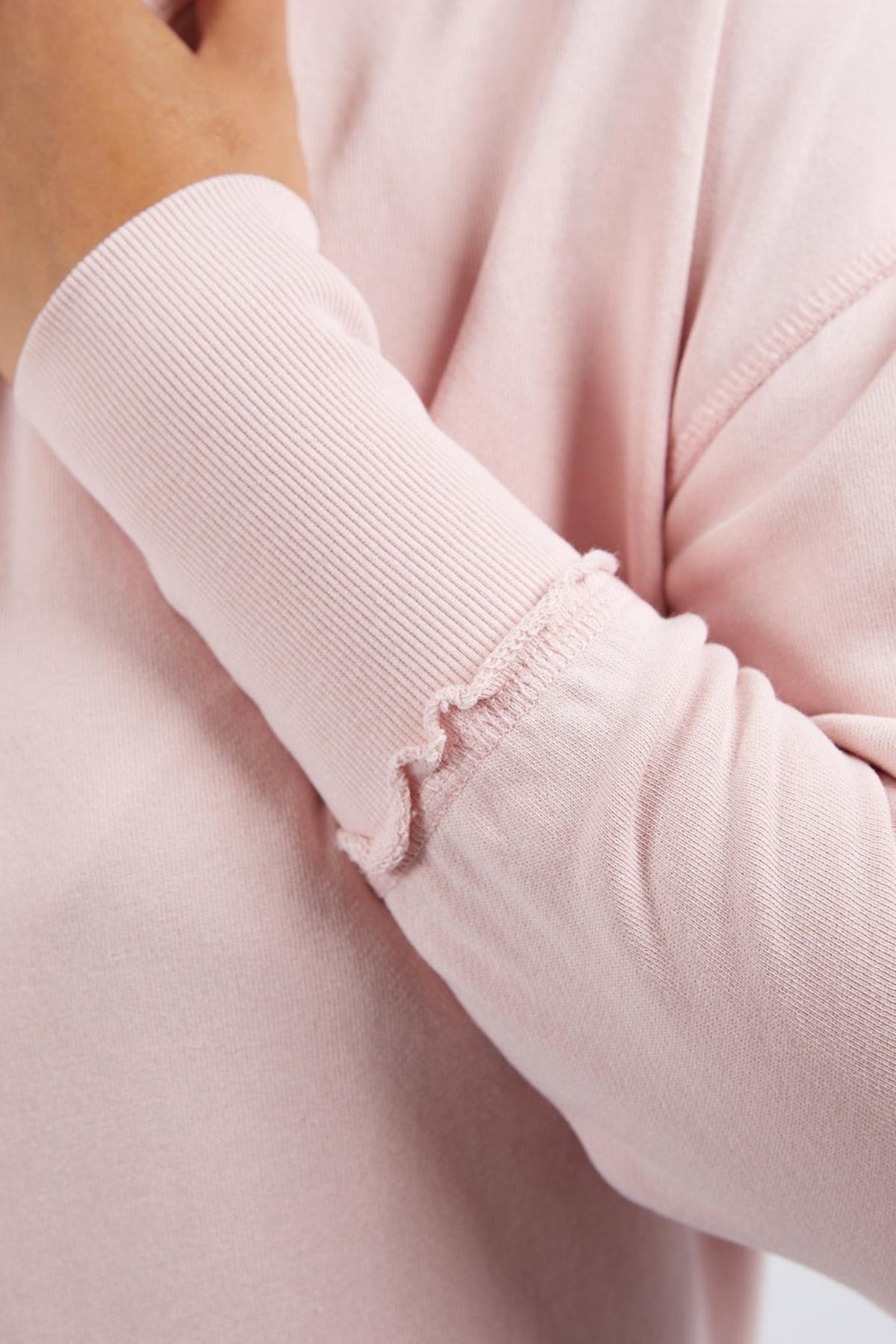 A close-up of a person wearing the light pink Foxwood Delilah Crew cotton jumper, which features ruffled sleeve edges. Their left hand is gently touching their neck near the collar.