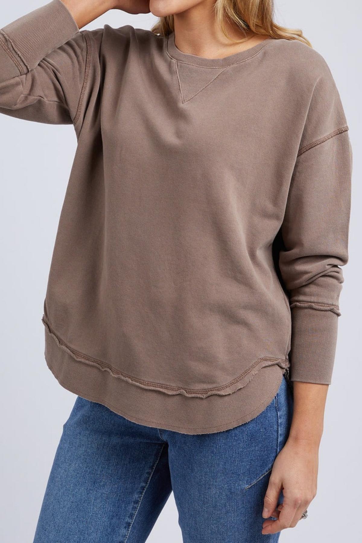 A woman models the Foxwood Delilah Crew, a loose taupe cotton jumper with exposed seams and crew neck, paired with blue jeans. She poses with one arm bent and hand near her head; only her torso and part of her face are visible.