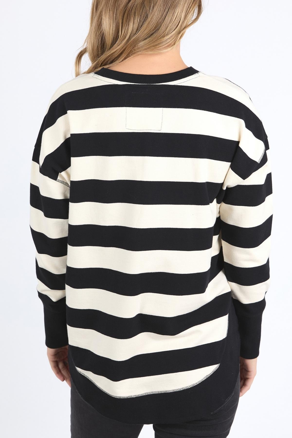 A person with long, blonde hair is seen from behind, wearing the Foxwood Delilah Stripe Crew—a black and white striped 100% cotton jumper—paired with black pants, against a plain white background.