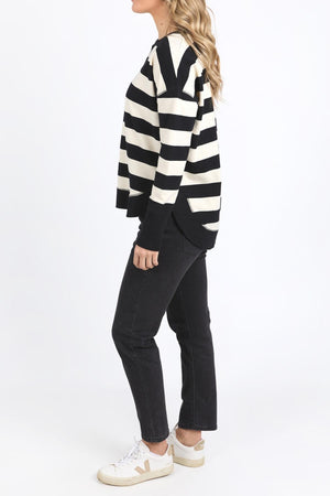 Woman standing sideways wearing a Foxwood Delilah Stripe Crew jumper, black jeans, and white sneakers with beige details. She has long wavy blond hair and is posed against a plain white background.