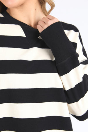 A person wears the Foxwood Delilah Stripe Crew, a black and white 100% cotton long-sleeve striped jumper. The image is cropped to show only the lower half of their face, upper body, and part of one arm near their face.