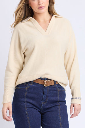 A woman wears the Foxwood Elea Knit Polo, a cream 100% cotton V-neck sweater with long sleeves, paired with blue high-waisted jeans and a brown belt, standing against a plain light background.