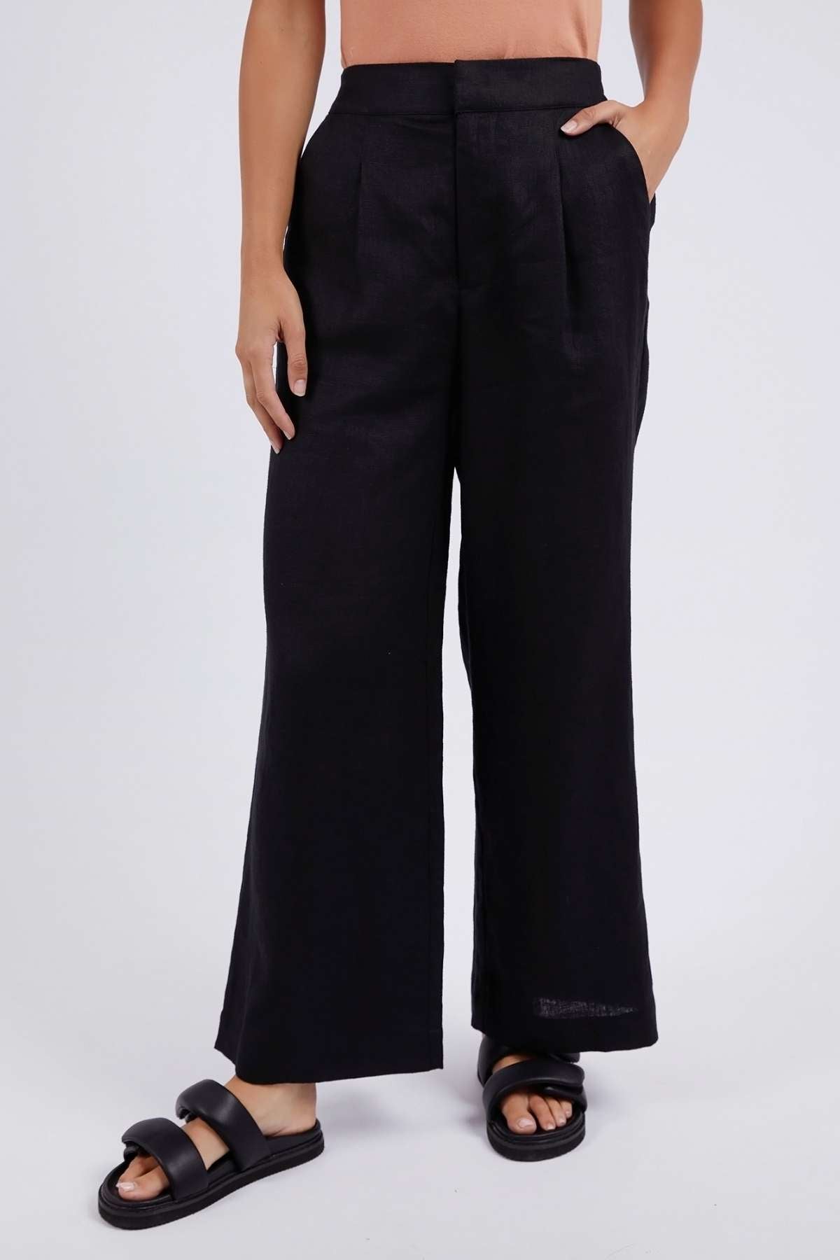 A person wears the Foxwood Elina Wide Leg Pant in black linen, styled relaxed and high-waisted with wide legs, hands in pockets and black slide sandals, standing against a plain light background. Their top and face are not shown.