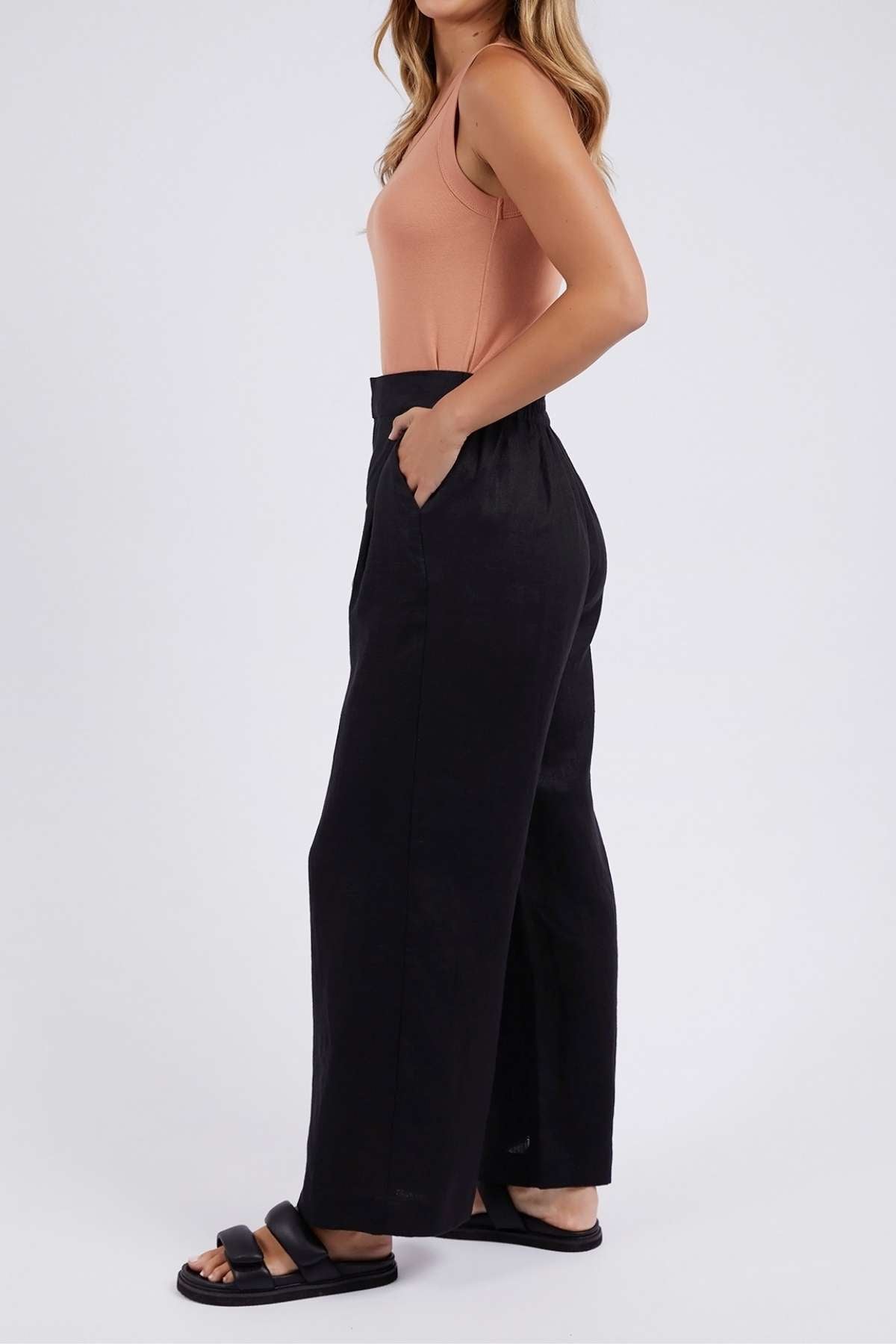 A woman stands in profile wearing a sleeveless peach top, Foxwood Elina Wide Leg Pant in black with a relaxed fit, and black slide sandals against a plain white background.