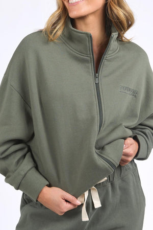 A woman smiles, holding the hem of her olive-green Foxwood Ember Zip Thru brushed cotton fleece jacket with a front chest logo, paired with matching pants featuring a white drawstring.