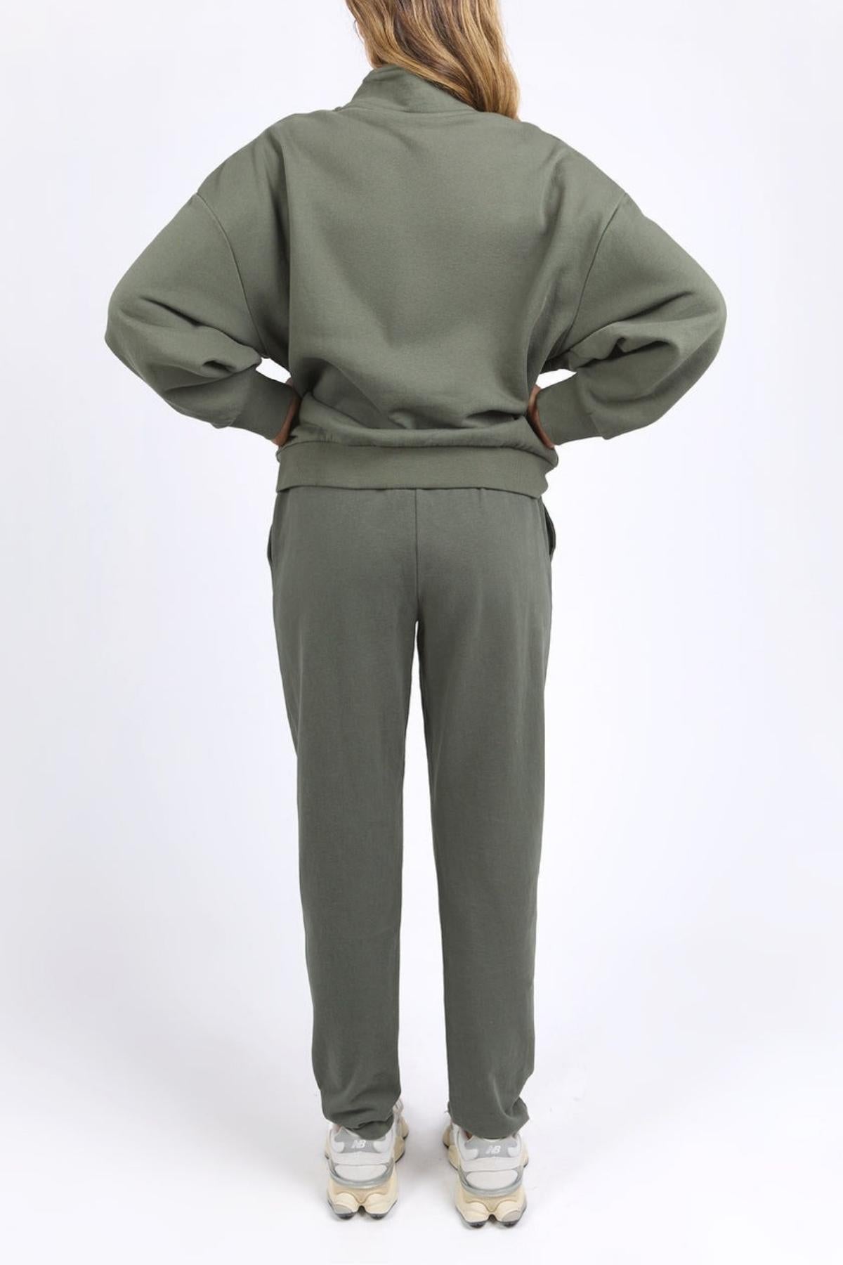 A person stands with hands on hips, facing away from the camera, wearing the Foxwood Ember Zip Thru in olive green and white sneakers against a plain white background.
