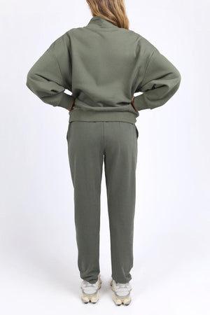 A person stands with hands on hips, facing away from the camera, wearing the Foxwood Ember Zip Thru in olive green and white sneakers against a plain white background.