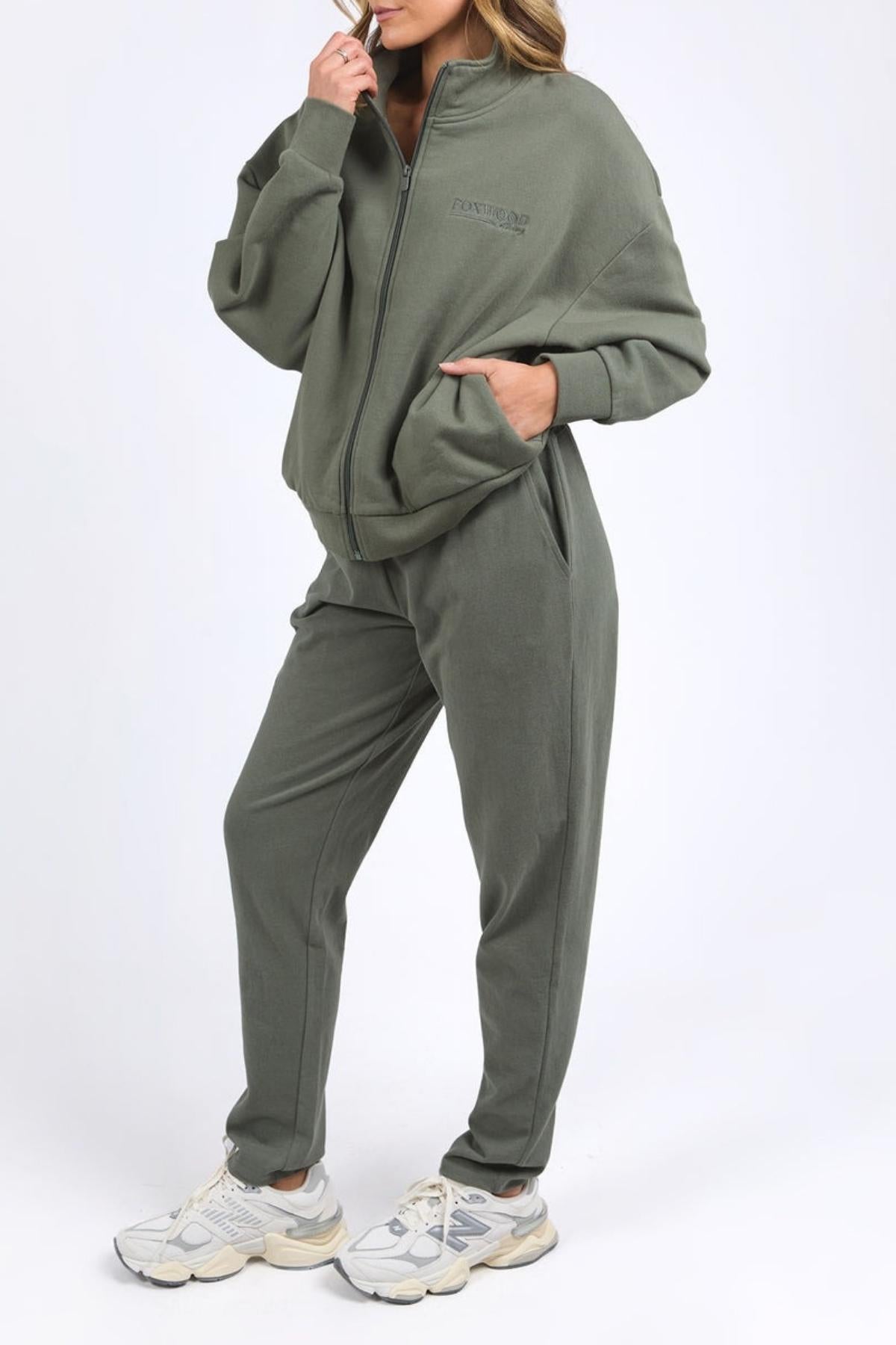 Wearing the Foxwood Ember Zip Thru, an olive green fleece jacket with a chest logo, a person stands against a plain white background with one hand in their pocket and the other touching their chin.