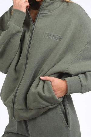 Wearing the olive green Foxwood Ember Zip Thru brushed cotton fleece jacket and matching pants, one hand in a pocket; the zip thru features a "FOXWOOD" front chest logo print.