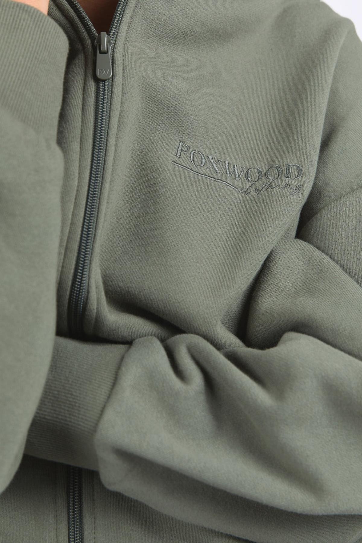 A close-up shows someone wearing the Foxwood Ember Zip Thru, a green brushed cotton fleece jacket with a chest logo print. Their arms are crossed, highlighting the matching green “FOXWOOD” embroidery on the sleeve.