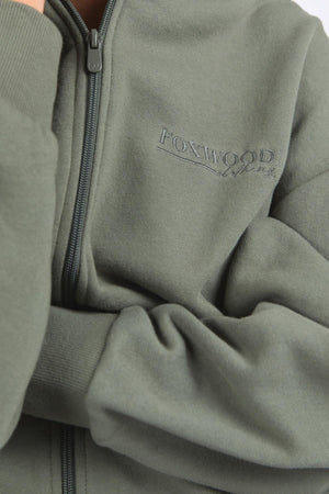 A close-up shows someone wearing the Foxwood Ember Zip Thru, a green brushed cotton fleece jacket with a chest logo print. Their arms are crossed, highlighting the matching green “FOXWOOD” embroidery on the sleeve.