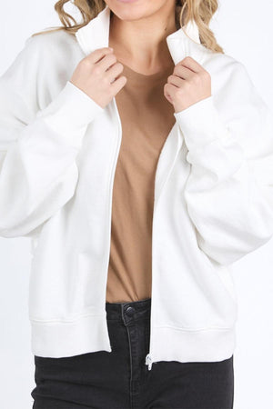 A person is shown from shoulders to hips holding the collar of a Foxwood Ember Zip Thru jacket, worn over a light brown T-shirt and black jeans. Their face is partially out of frame.