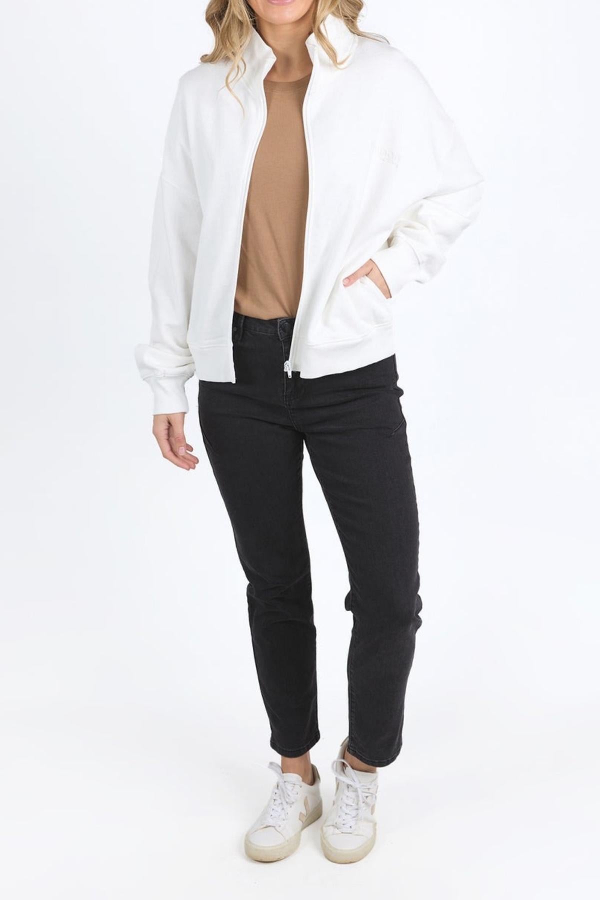Wearing the Foxwood Ember Zip Thru brushed cotton fleece jacket with chest logo over a tan shirt, black jeans, and white sneakers, the person stands against a plain white background with hands in pockets and face partly out of frame.