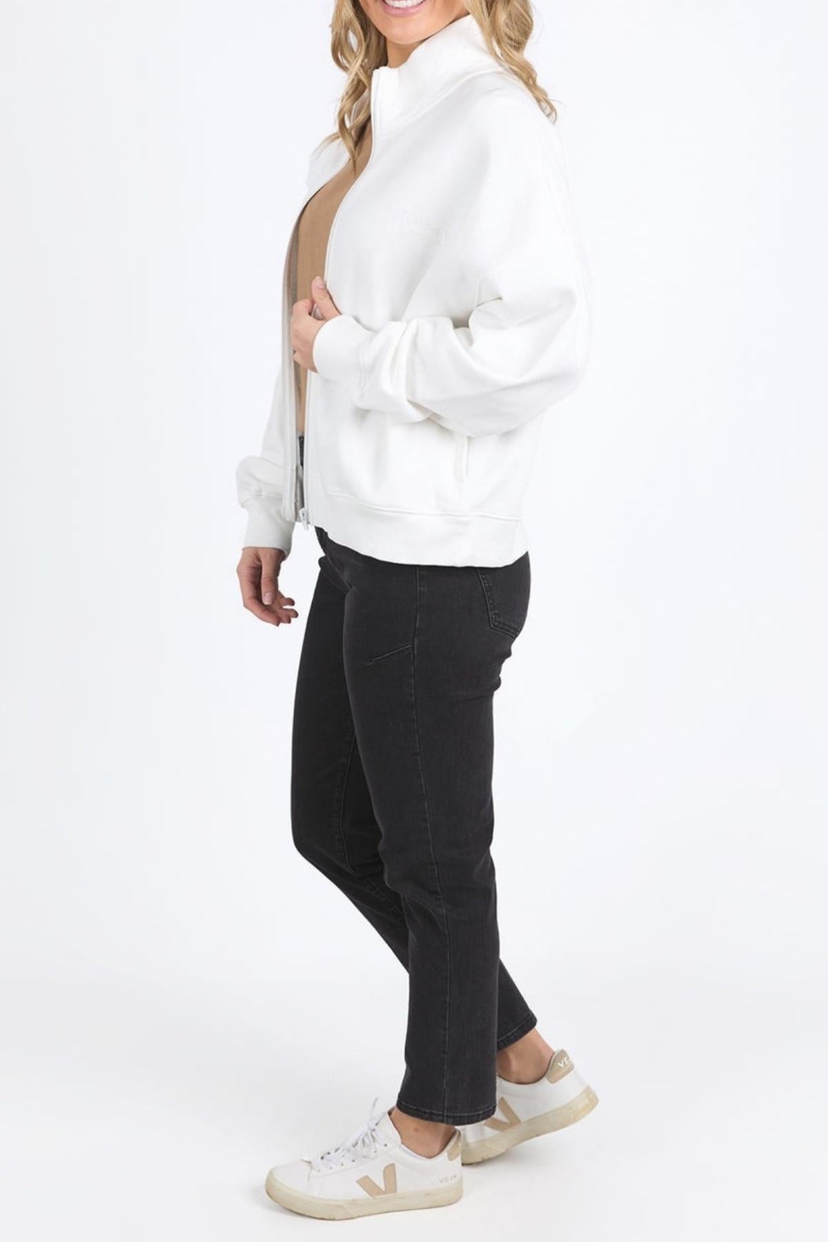 A woman stands sideways wearing the Foxwood Ember Zip Thru white brushed cotton fleece jacket with a chest logo, beige top, black jeans, and white sneakers with beige accents against a plain white background.