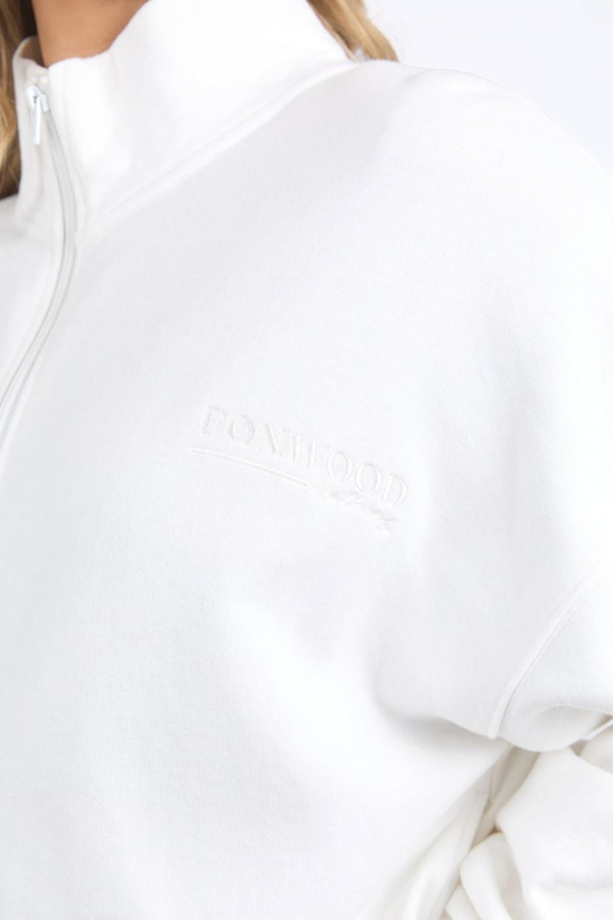 Close-up of someone wearing the Foxwood Ember Zip Thru brushed cotton fleece jacket, featuring an embroidered chest logo on a white sweatshirt, shown against a plain light background.