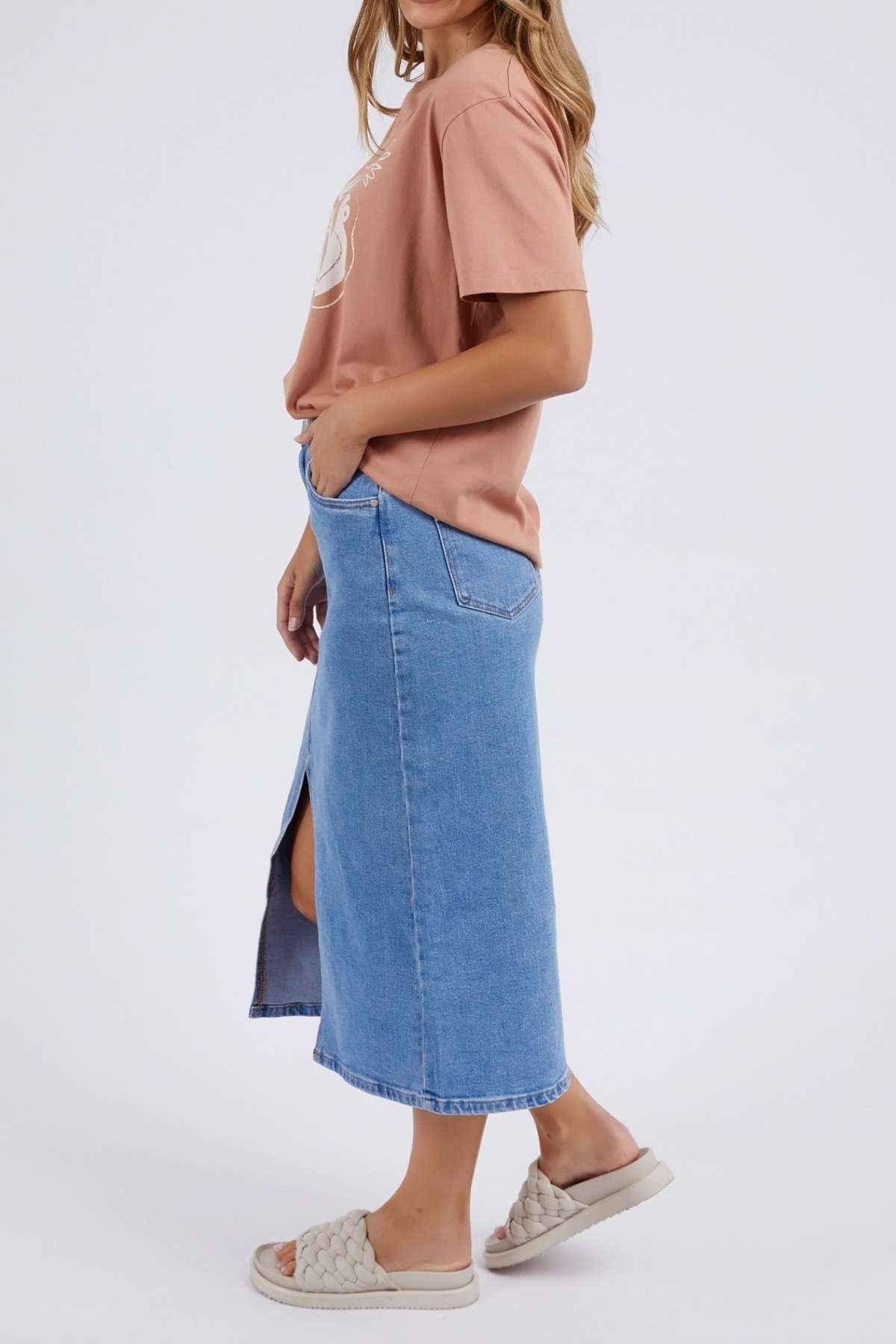 A woman in a relaxed-fit, light pink Foxwood Etched Geo Tee and a blue denim skirt with side slit stands sideways, hands in pockets, wearing quilted slide sandals against a plain white background.