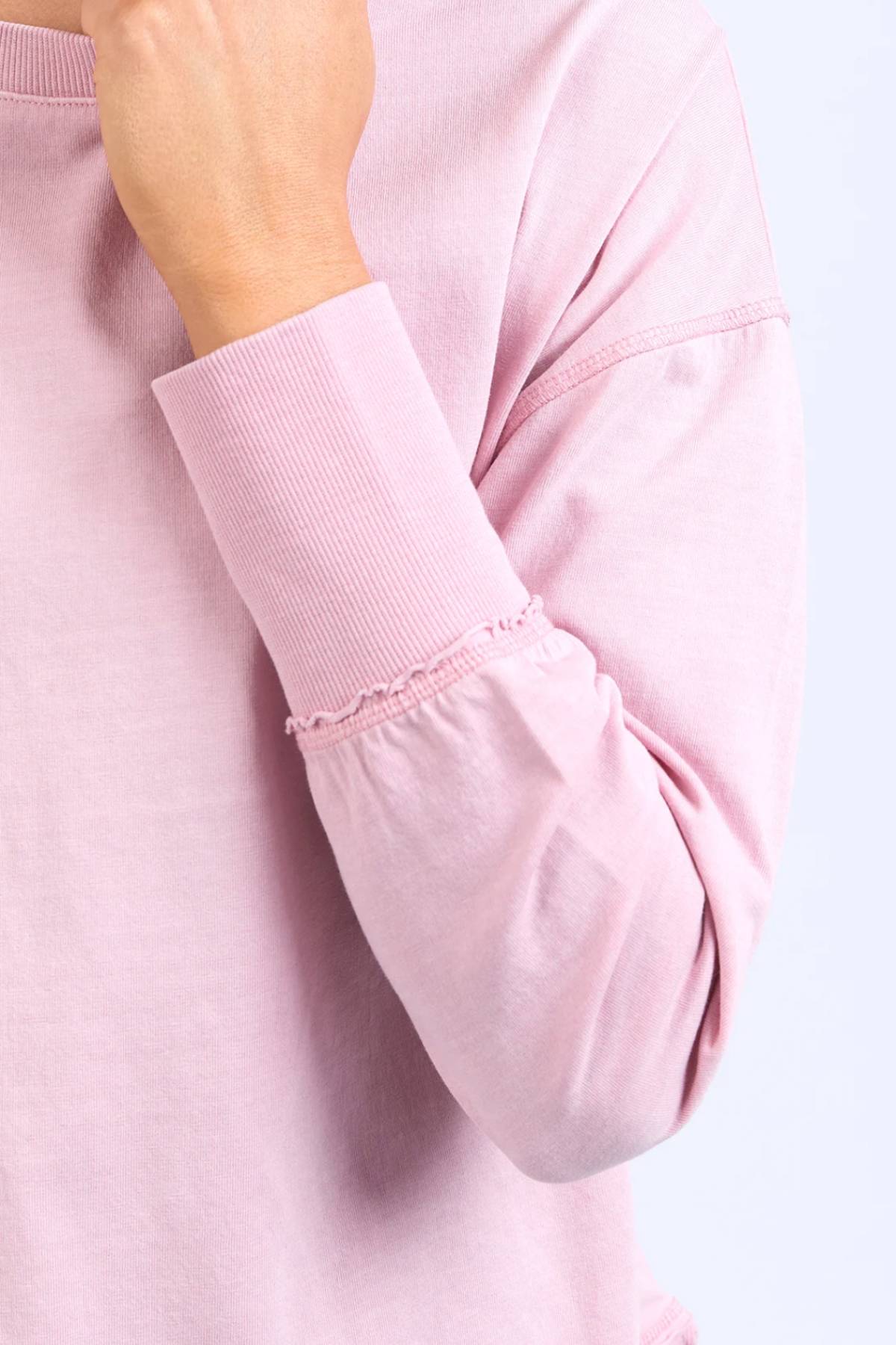 A person in the Foxwood Farrah Long Sleeve, a relaxed fit light pink top with ribbed cuffs and subtle stitching, holds their hand near their face against a light background.