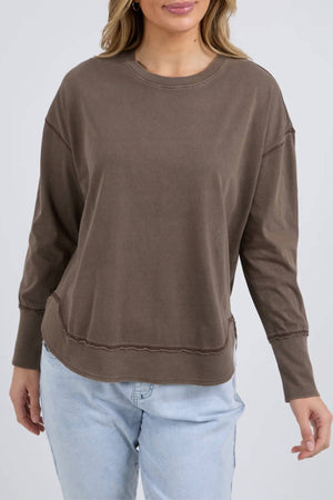 A woman wears the Foxwood Farrah Long Sleeve, a relaxed fit brown top, paired with light blue jeans against a neutral background. Her face is partially out of frame.