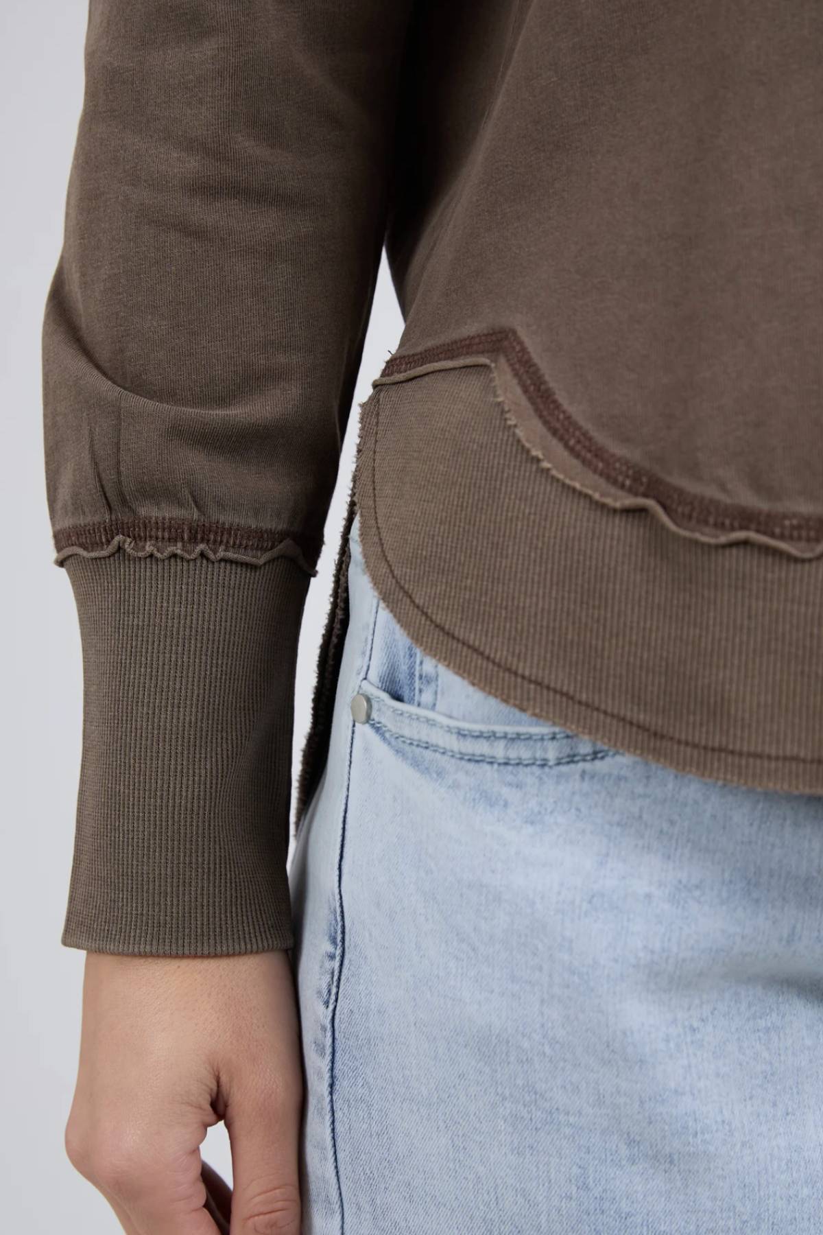 A close-up of a person in the Foxwood Farrah Long Sleeve, a brown relaxed fit top with scalloped hem, paired with light blue jeans; left arm and side of torso are visible.