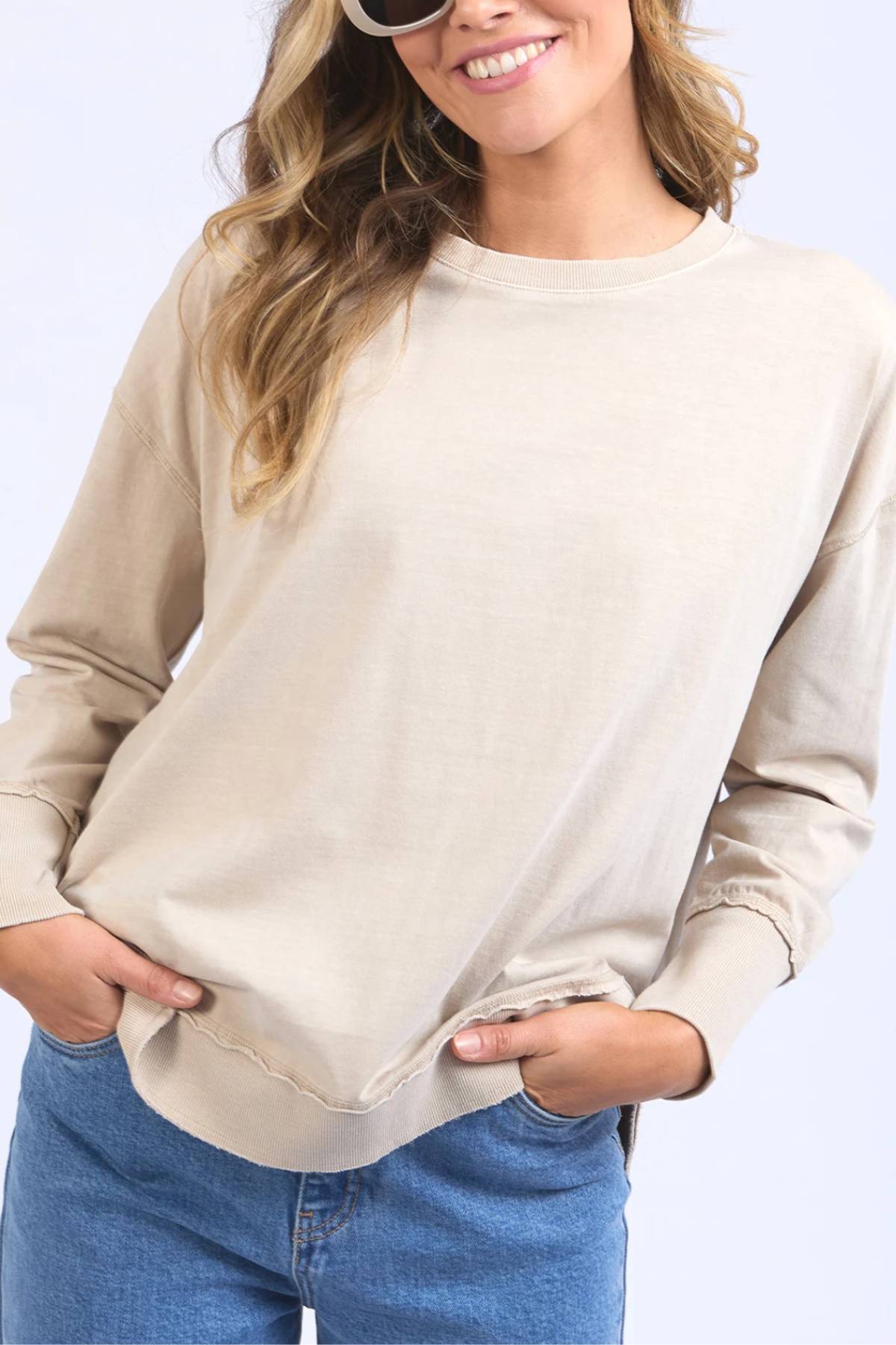 A woman in sunglasses, blue jeans, and a beige Foxwood Farrah Long Sleeve top smiles with her hands in her pockets against a light background.
