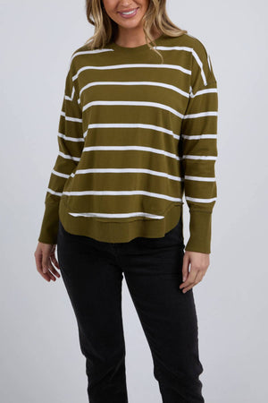 A woman wearing the Foxwood Farrah Stripe Long Sleeve in olive green with white stripes, made from 100% Fresh Pima Cotton, and black pants stands smiling against a plain light background with one hand by her side.