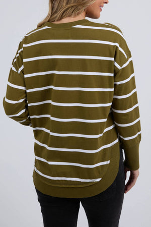A person with light brown hair is shown from the back, wearing the Foxwood Farrah Stripe Long Sleeve jumper in olive and white, made from 100% Fresh Pima Cotton, featuring a rounded hem, long sleeves, and paired with black pants.