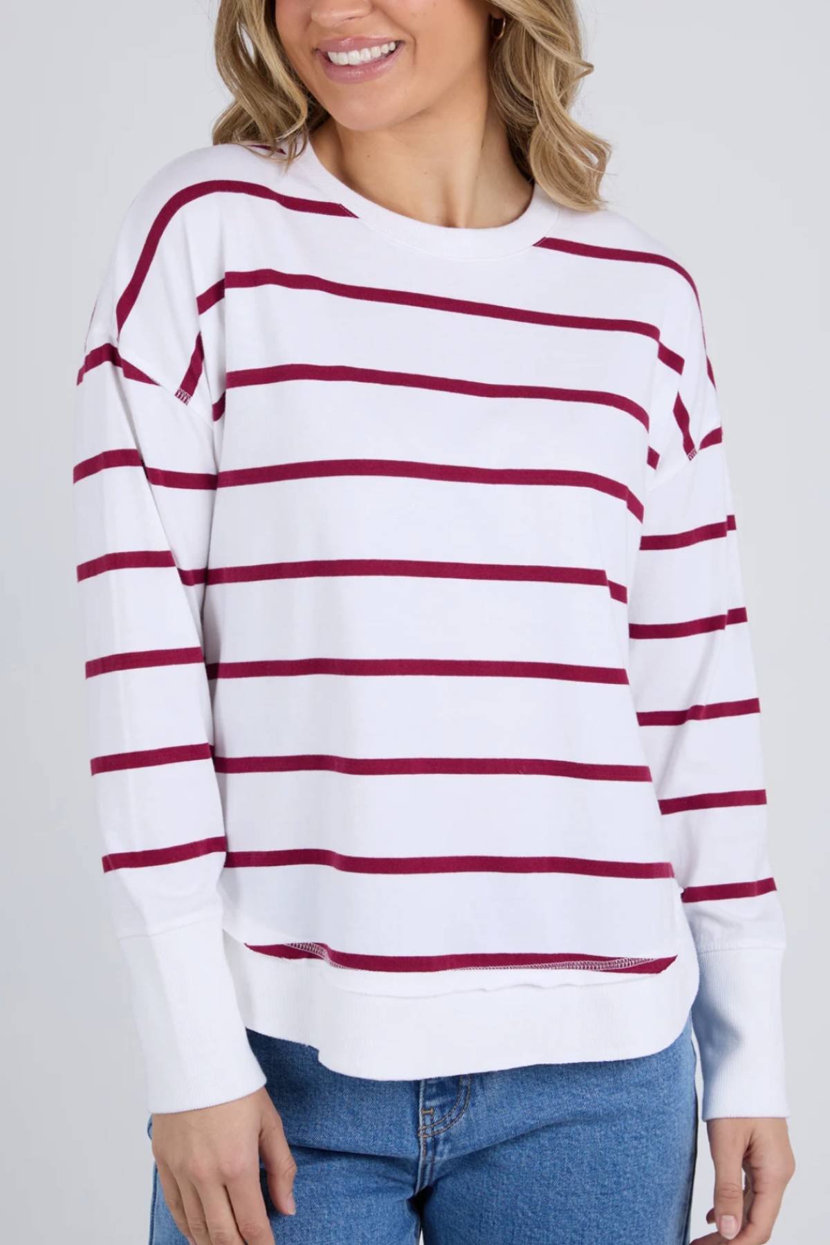 A woman smiles against a plain light background wearing the Foxwood Farrah Stripe Long Sleeve, featuring white fabric with horizontal maroon stripes and paired with blue jeans.