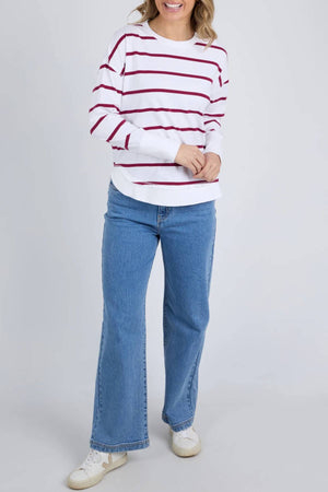 Wearing a Foxwood Farrah Stripe Long Sleeve with blue jeans and white sneakers, a person stands against a plain light background, smiling with hands clasped in front.